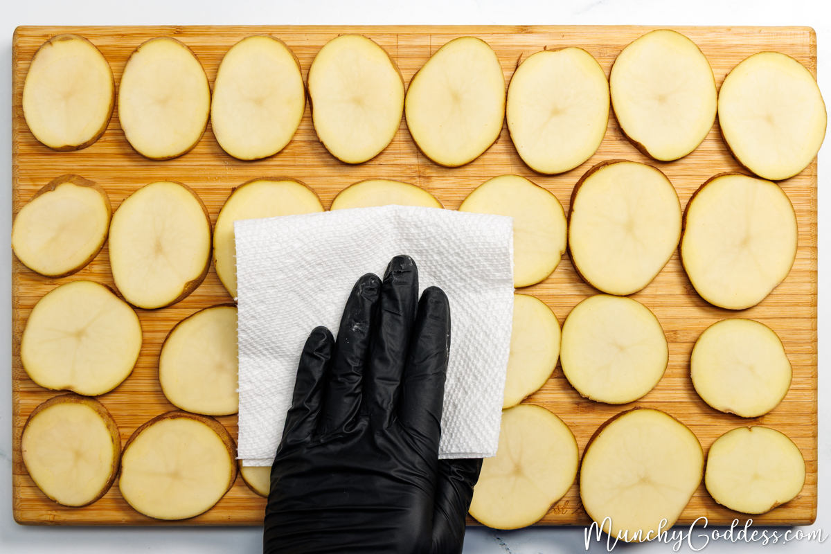Potato slices spread out on a wood cutting board being patted dry with a paper towel.