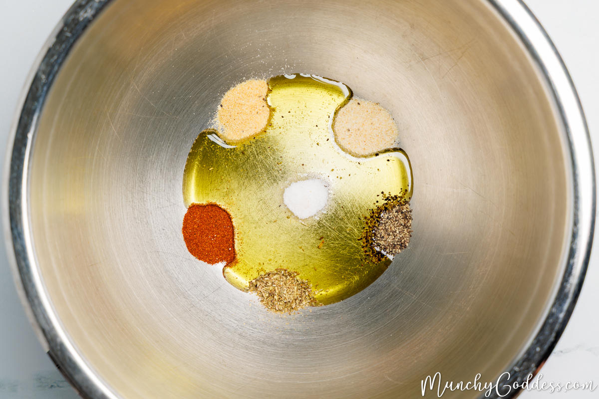 Olive oil, salt, ground black pepper, garlic powder, onion powder, smoked paprika, and Italian seasoning added to a silver mixing bowl.