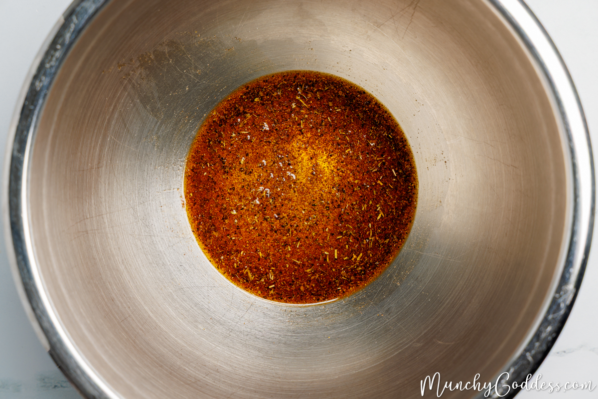 Olive oil, salt, ground black pepper, garlic powder, onion powder, smoked paprika, and Italian seasoning well blended into an oil seasoning mixture in a silver mixing bowl.