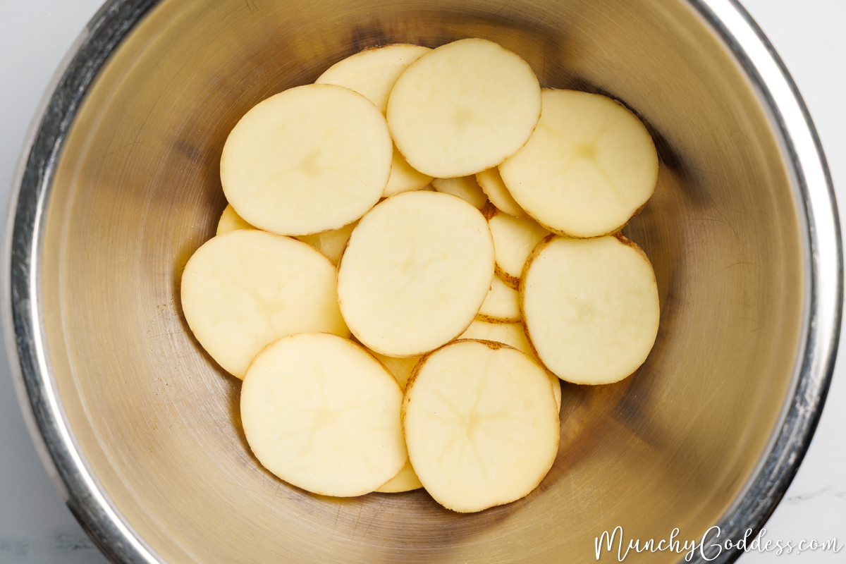 Potato slices added to an oil seasoning mixture in a silver mixing bowl.