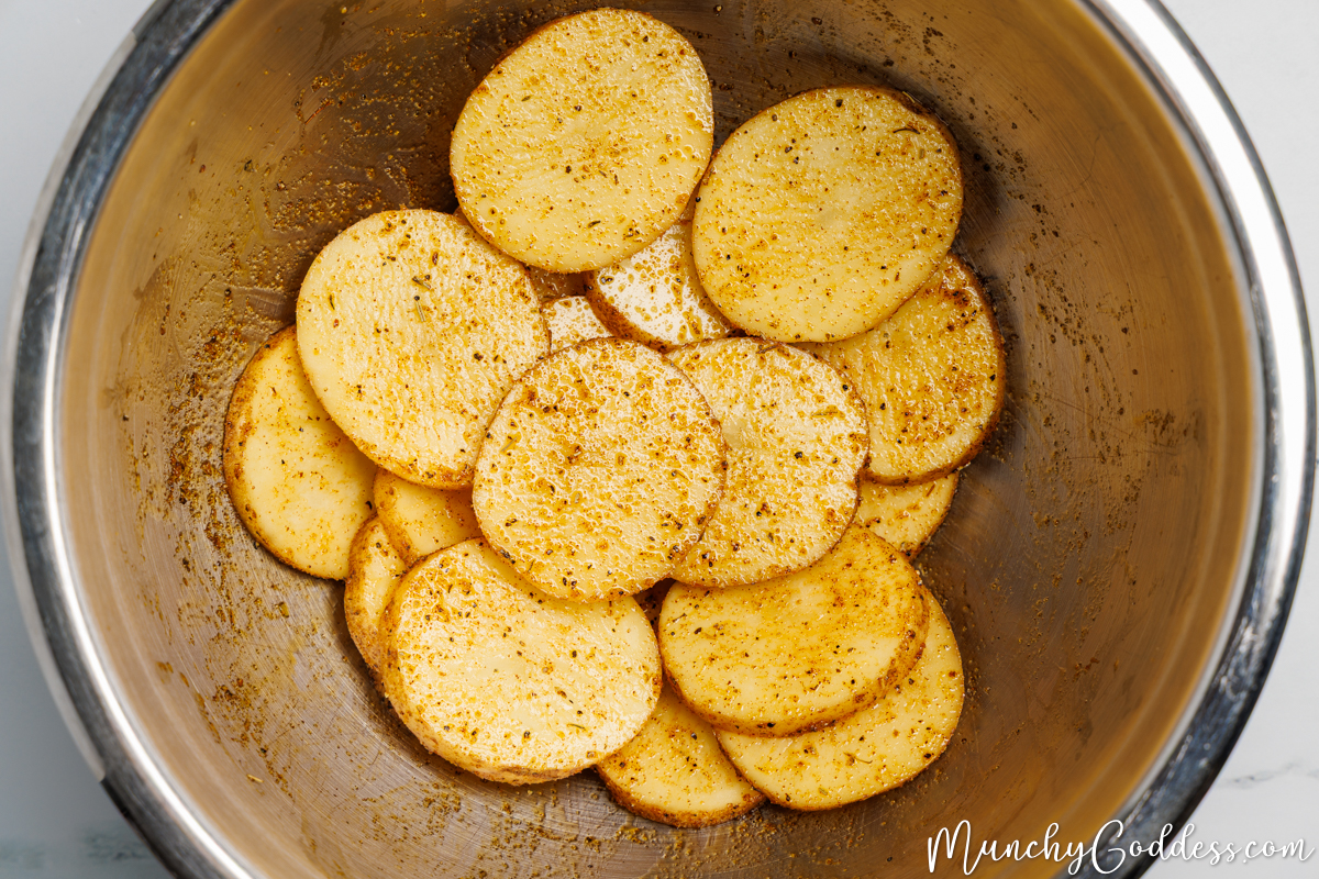 Potato slices tossed until well coated in an oil seasoning mixture in a silver mixing bowl.