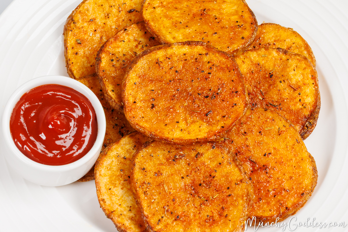 Oven baked potato slices and a sauce dish filled with ketchup on a round white plate.