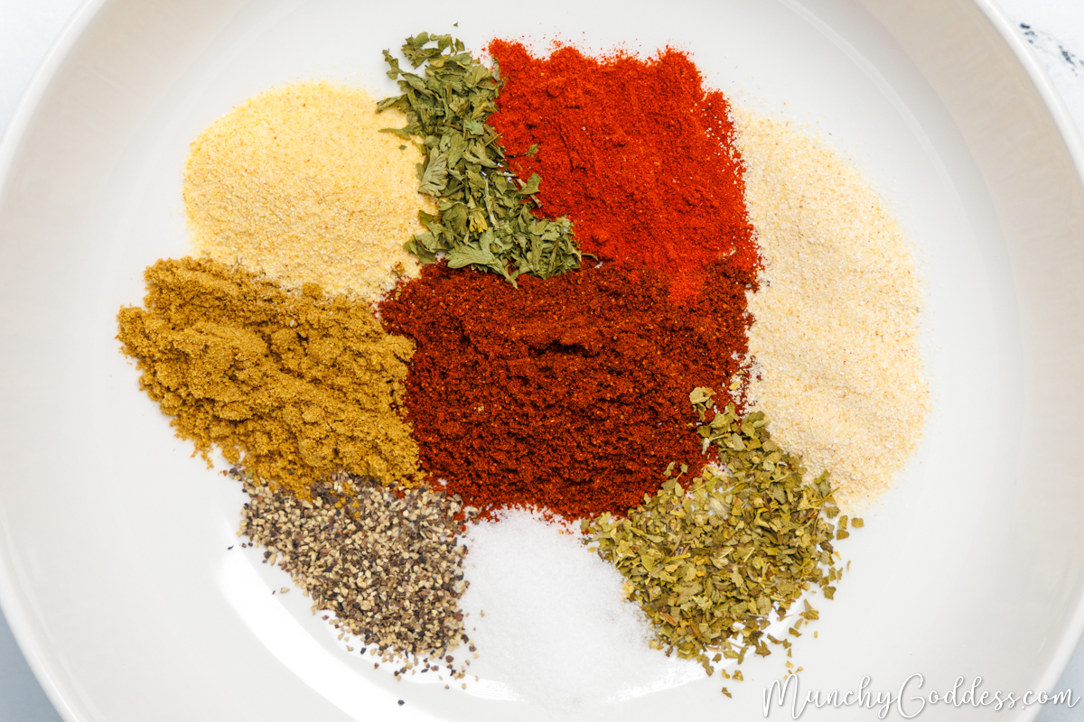Smoked paprika, ground cumin, garlic powder, onion powder, dried oregano, salt, chili powder, parsley flakes, and ground black pepper neatly placed in a white bowl.