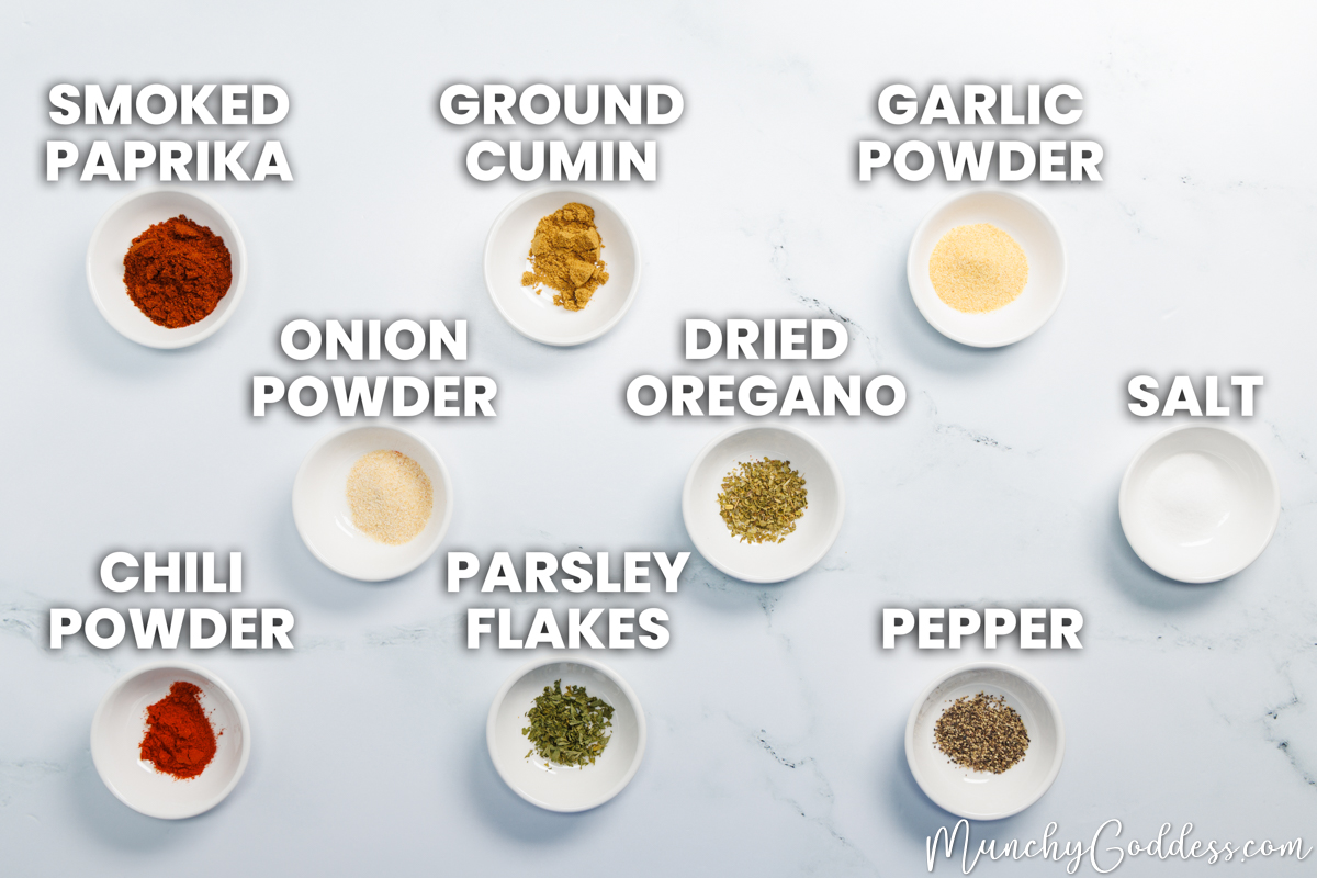 Taco seasoning recipe ingredients in individual ceramic dishes on a white marble surface: smoked paprika, ground cumin, garlic powder, onion powder, dried oregano, salt, chili powder, parsley flakes, and ground black pepper.