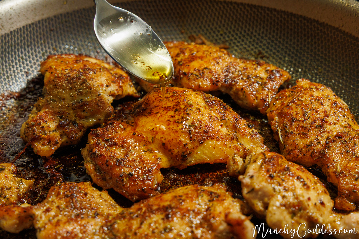 A matte silver spoon drizzling butter sauce on top of a cooked cajun butter chicken thigh in a skillet.
