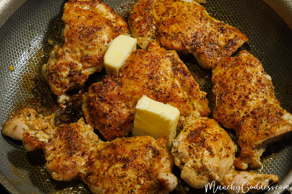 Two pieces of butter in between Cajun seasoned chicken thighs that have been cooking in a skillet.