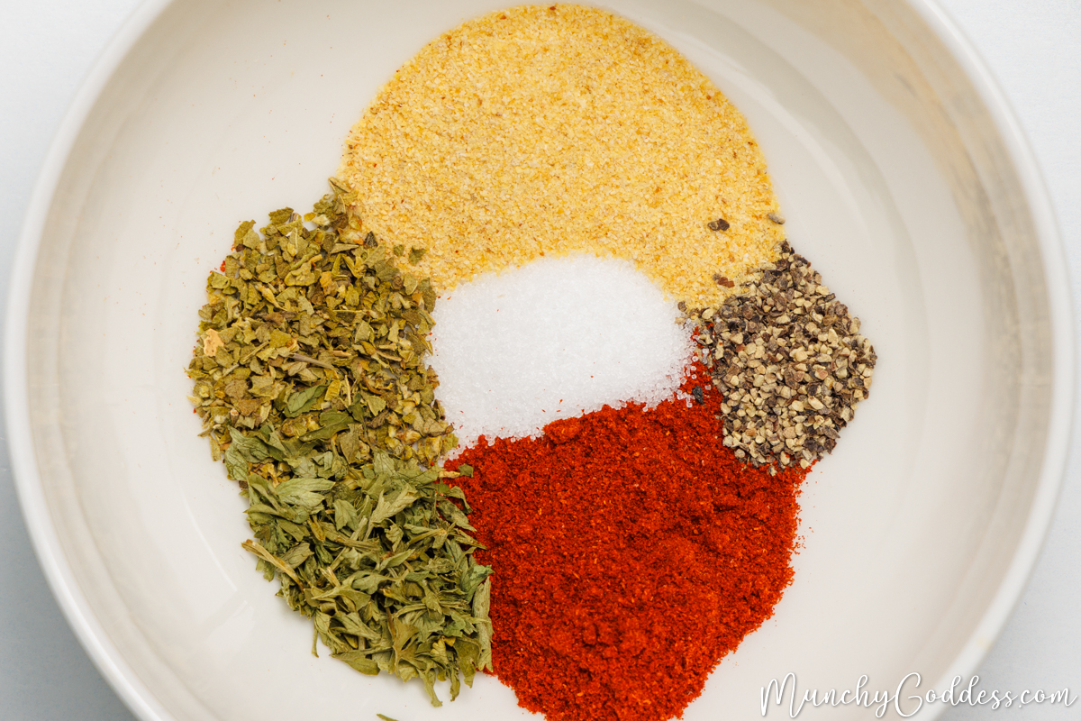 Dried oregano, parsley flakes, smoked paprika, salt, ground black pepper, and garlic powder neatly placed in an off white bowl.