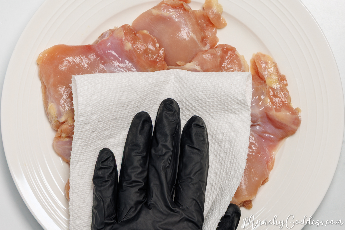 Boneless skinless chicken thighs on an off white plate being patted dry with a paper towel.