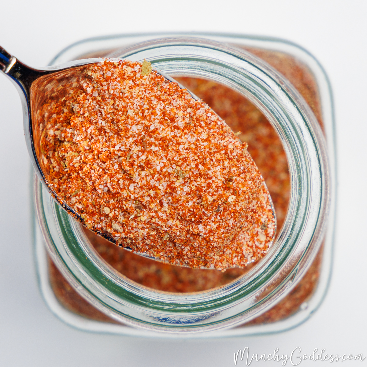 A scoop of Cajun seasoning resting on top of a clear glass spice jar filled with Cajun seasoning.