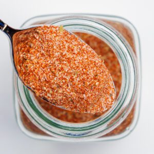 A scoop of Cajun seasoning resting on top of a clear glass spice jar filled with Cajun seasoning.