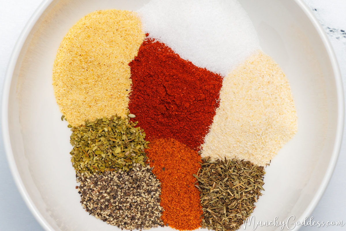 Smoked paprika, salt, onion powder, dried thyme, cayenne pepper, ground black pepper, dried oregano, and garlic powder neatly placed in an off-white bowl.