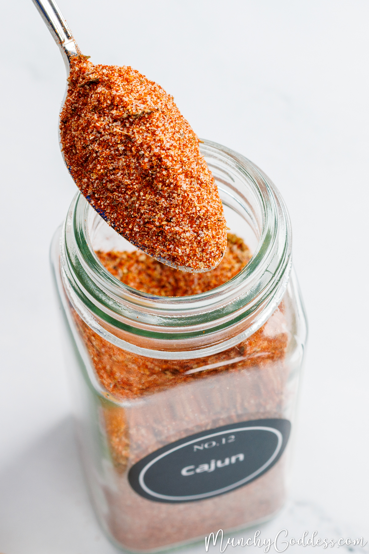 A small spoon removing a scoop of Cajun seasoning from a clear glass spice jar with a black label that says "Cajun".