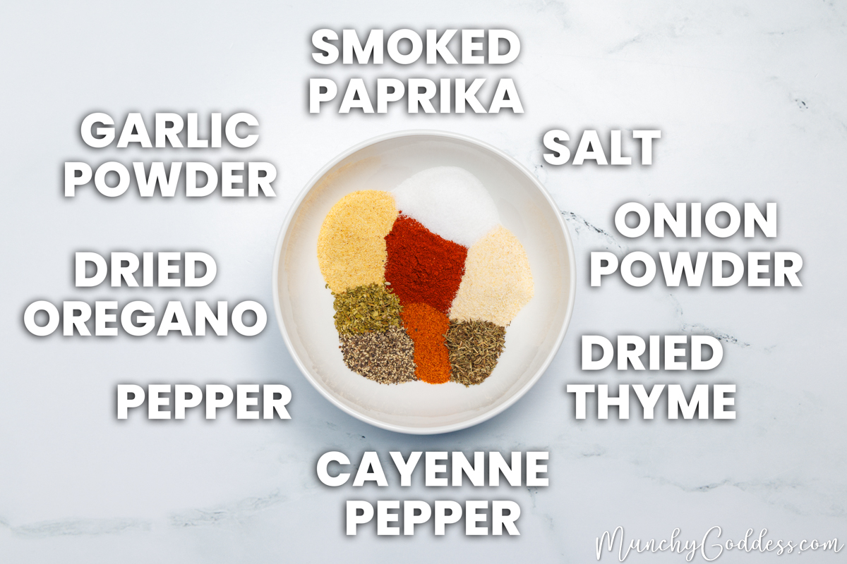Cajun seasoning recipe ingredients placed neatly inside a bowl on a white marble surface: smoked paprika, salt, onion powder, dried thyme, cayenne pepper, ground black pepper, dried oregano, and garlic powder.