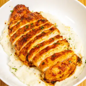 Sliced juicy air fryer chicken breast on a bed of mashed potatoes topped with freshly chopped parsley in an off-white bowl.