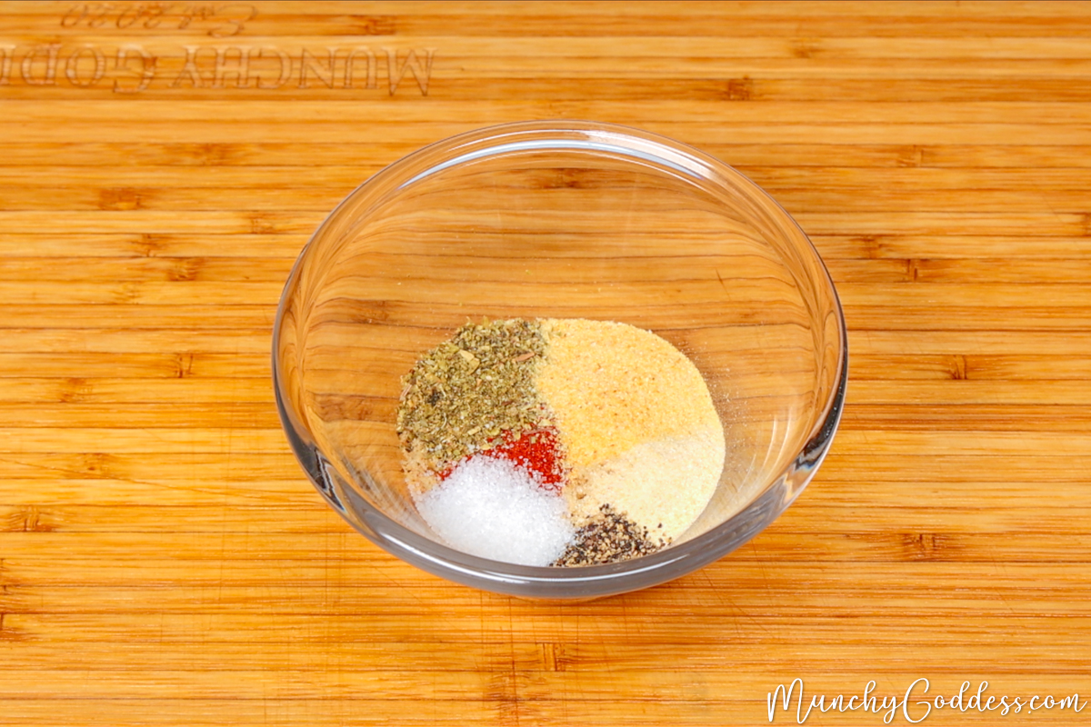 Brown sugar, smoked paprika, Italian seasoning, garlic powder, onion powder, salt, and ground black pepper neatly placed in a small glass bowl on a wood cutting board.