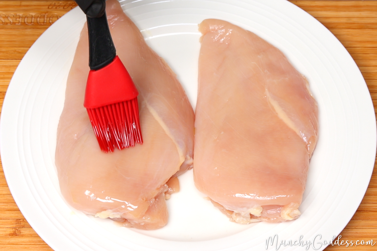 Olive oil being brushed on a chicken breast with a red and black basting brush on an off-white plate.