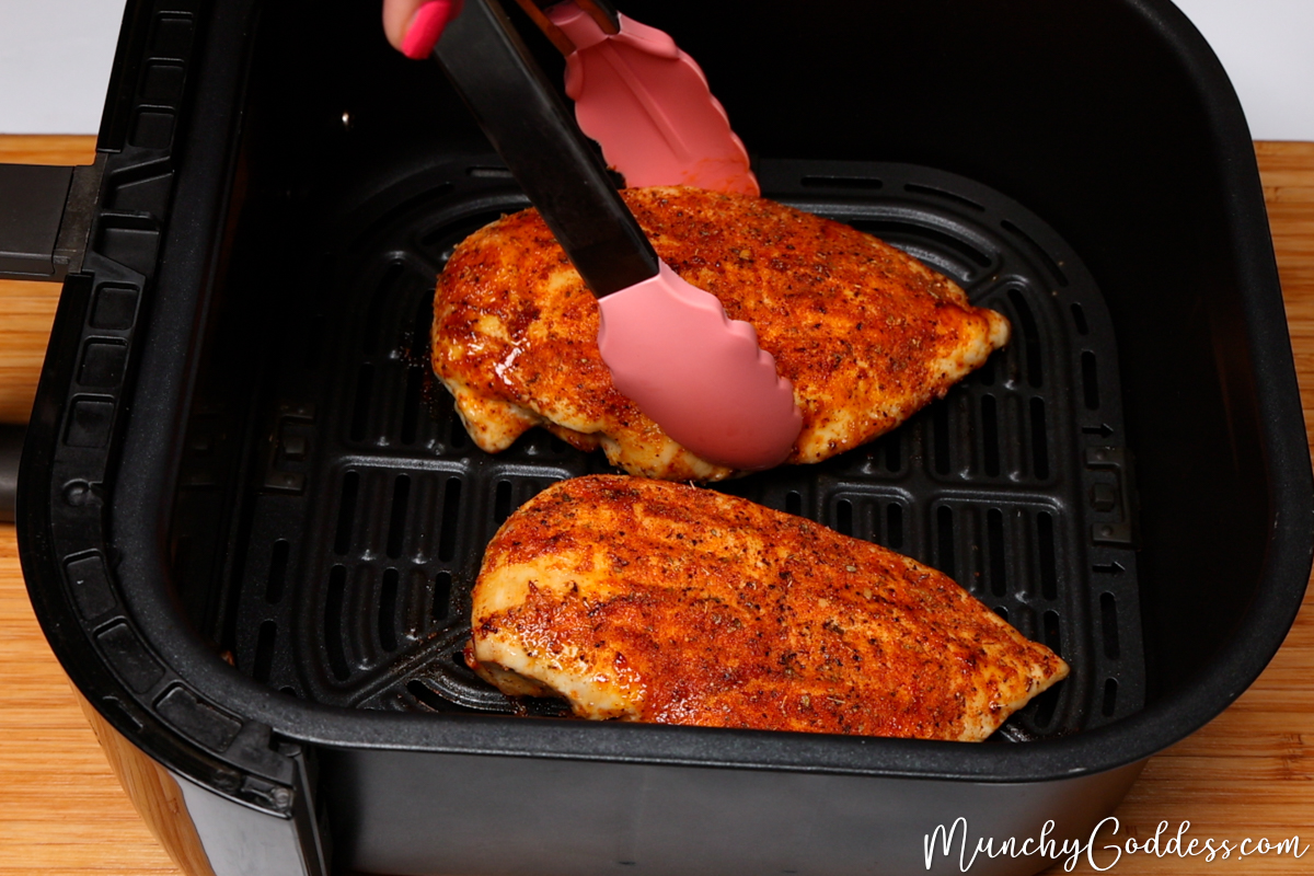Chicken breast being flipped in a black air fryer basket with pink and silver tongs after 10 minutes of cooking.