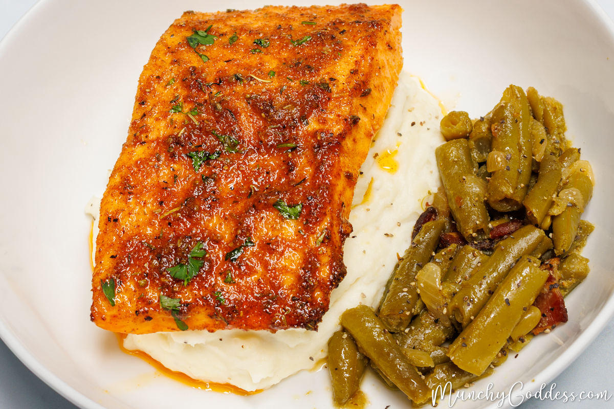 Cooked salmon fillet on top of mashed potatoes next to a side of green beans in an off-white bowl.