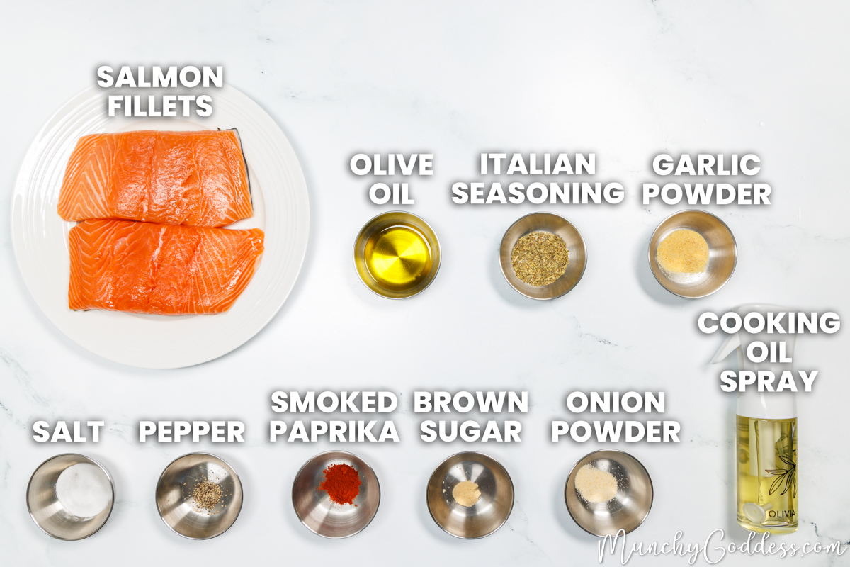 Air Fryer Salmon recipe ingredients in various dishes on a white marble surface: salmon filets, olive oil, Italian seasoning, garlic powder, salt, ground black pepper, smoked paprika, brown sugar, onion powder, cooking oil spray.