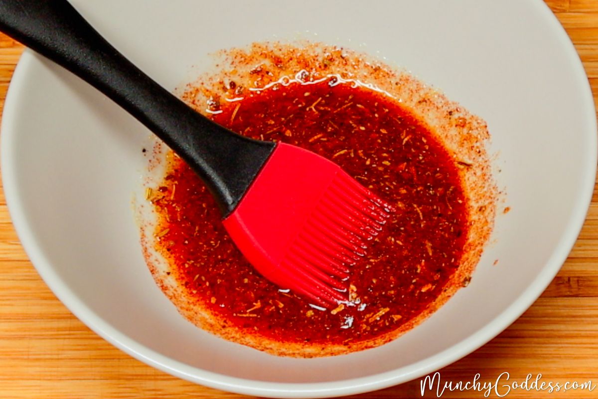 Olive oil, Italian seasoning, garlic powder, salt, ground black pepper, smoked paprika, brown sugar, and onion powder blended into an olive oil seasoning mixture with a red and black basting brush in a light gray bowl.