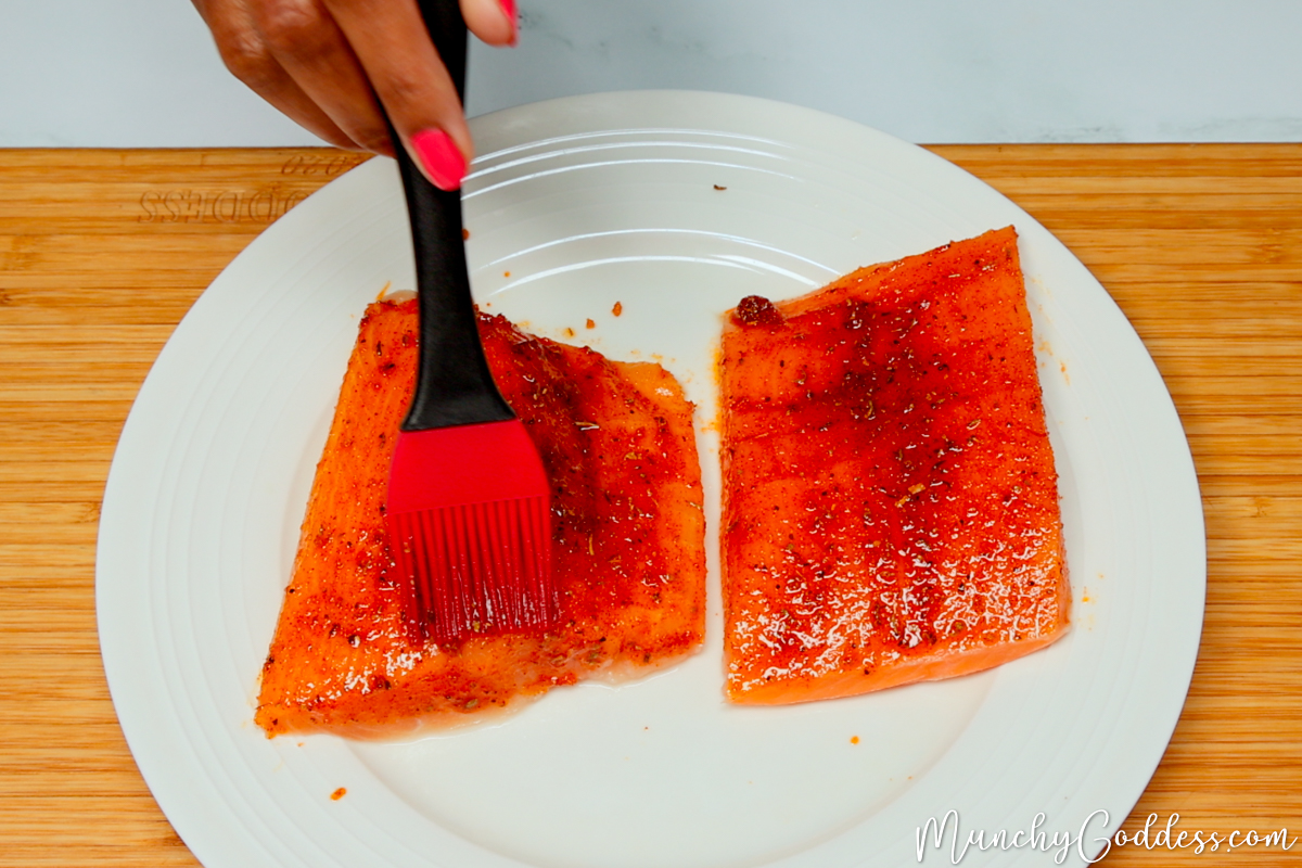 Salmon fillets being coated with an olive oil seasoning mixture with a red and black basting brush on an off-white plate.