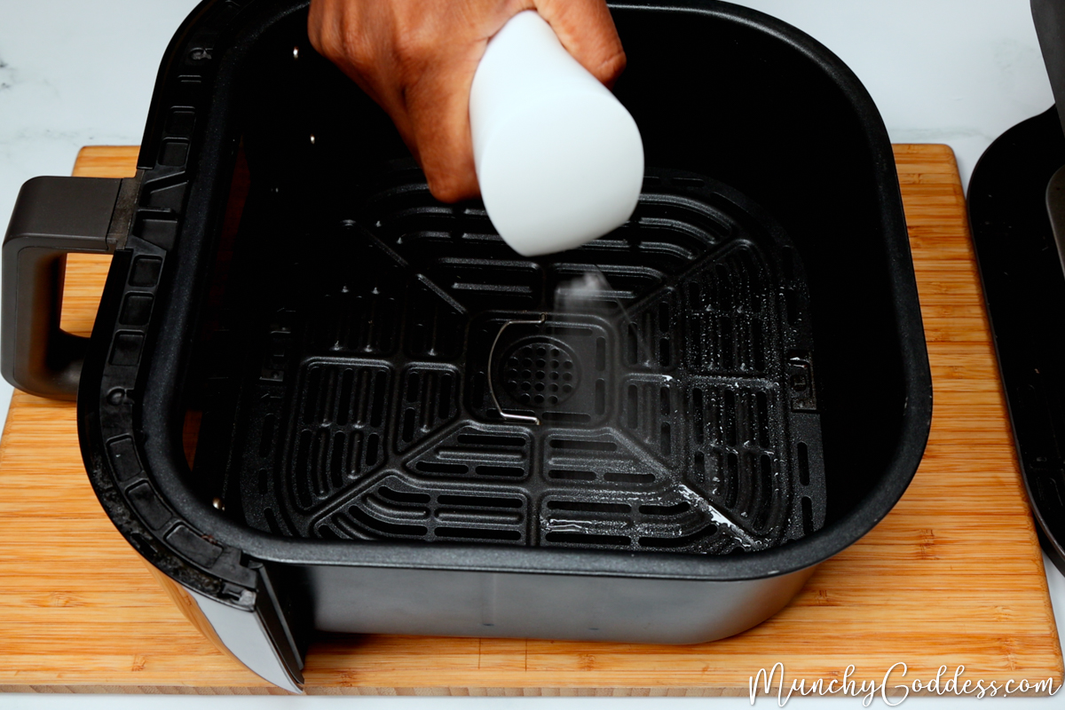 Cooking oil being sprayed onto an air fryer tray inside an air fryer basket.