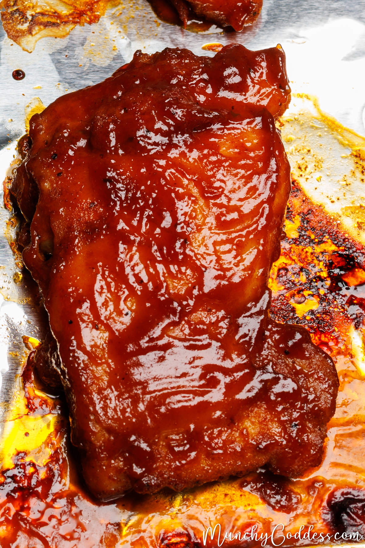 Baked BBQ chicken thigh with crispy edges glistening with BBQ sauce on a sheet pan lined with foil.