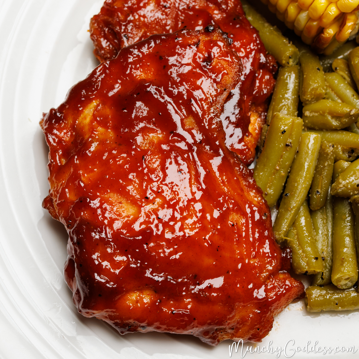 Baked BBQ chicken thighs on an off-white plate next to sides of green beans and corn on the cob.
