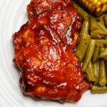Baked BBQ chicken thighs on an off-white plate next to sides of green beans and corn on the cob.