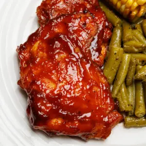 Baked BBQ chicken thighs on an off-white plate next to sides of green beans and corn on the cob.