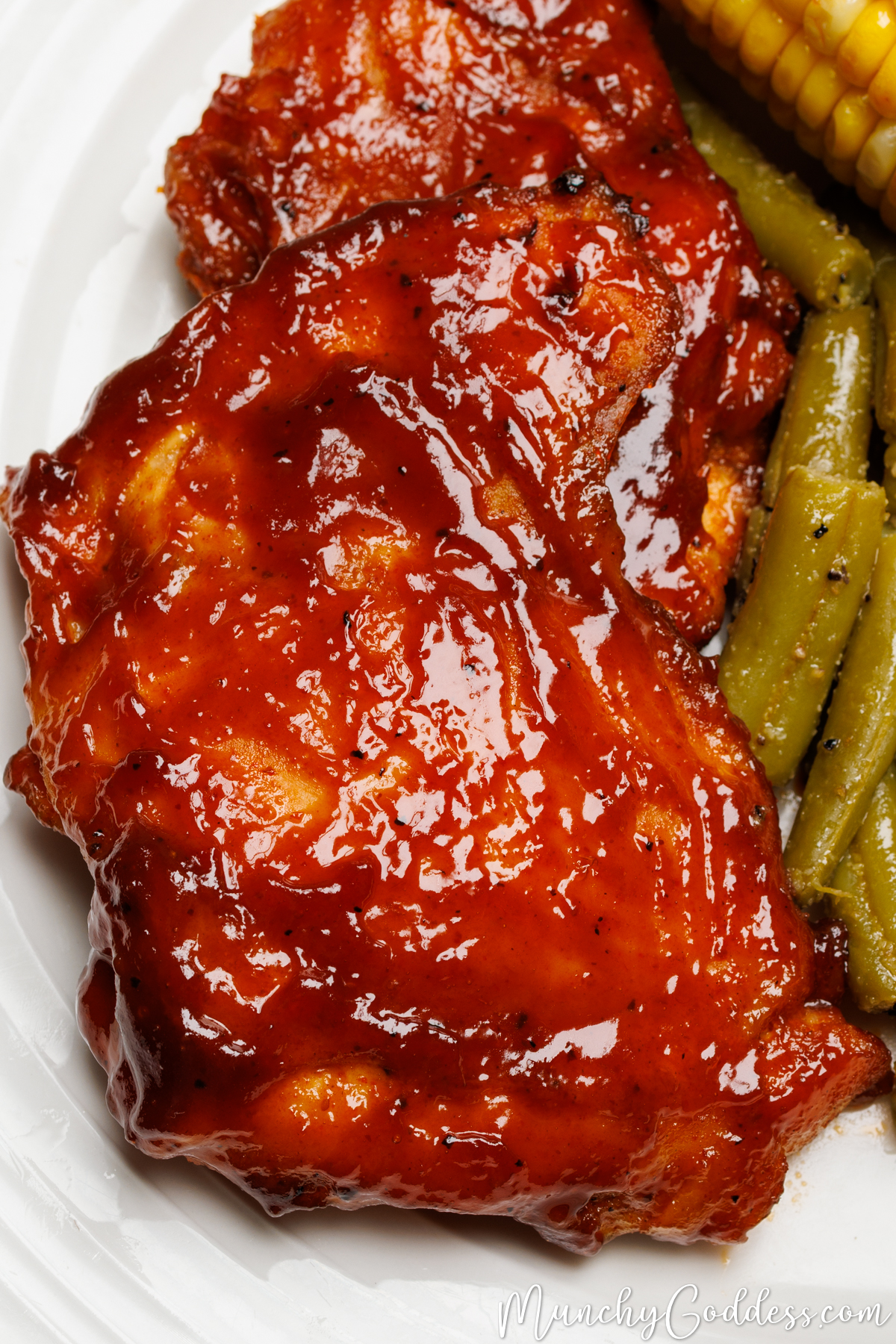 Baked BBQ chicken thighs on an off-white plate next to green beans and corn on the cob.