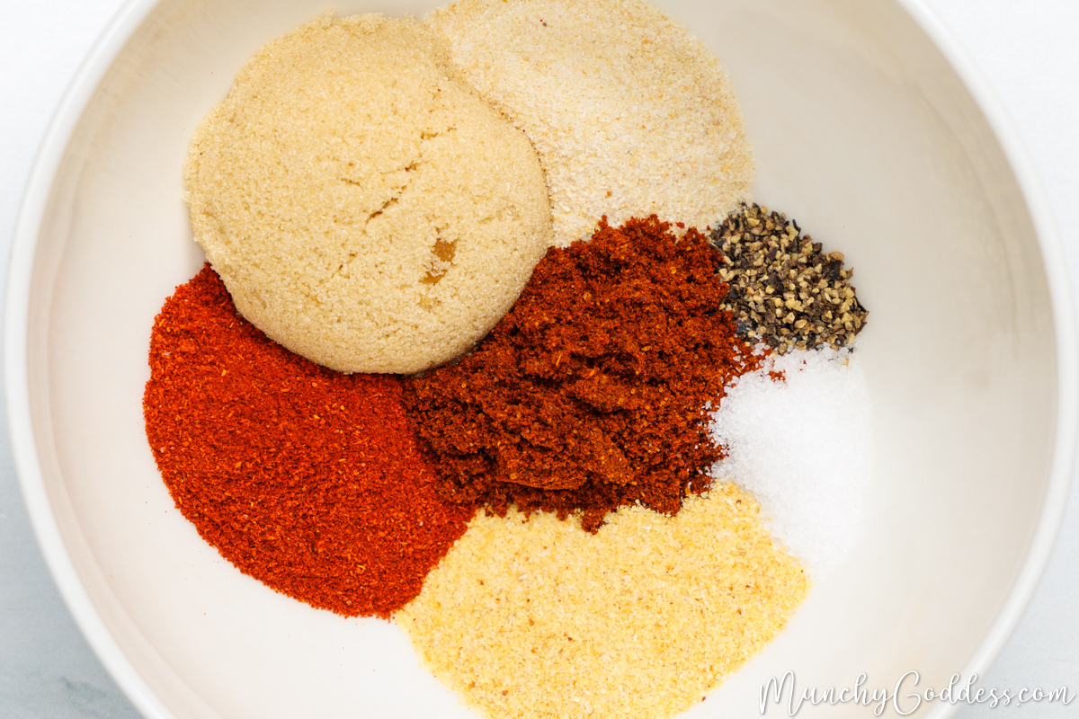 Brown sugar, onion powder, ground black pepper, salt, garlic powder, smoked paprika, and chili powder neatly placed in an off-white bowl.