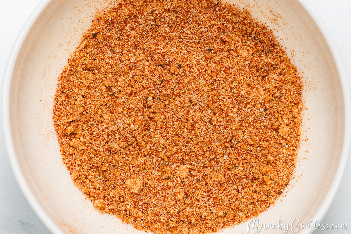 Brown sugar, onion powder, ground black pepper, salt, garlic powder, smoked paprika, and chili powder blended to become a seasoning in an off-white bowl.