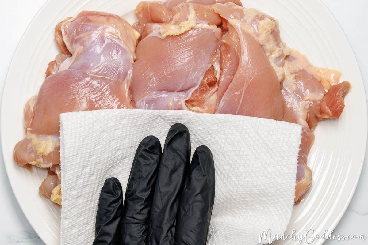 Boneless skinless chicken thighs on an off-white plate being patted dry with a paper towel.