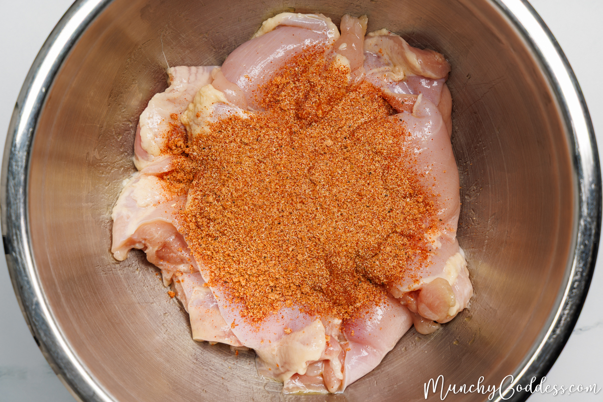A seasoning mixture sprinkled on top of boneless skinless chicken thighs coated in olive oil in a silver mixing bowl.