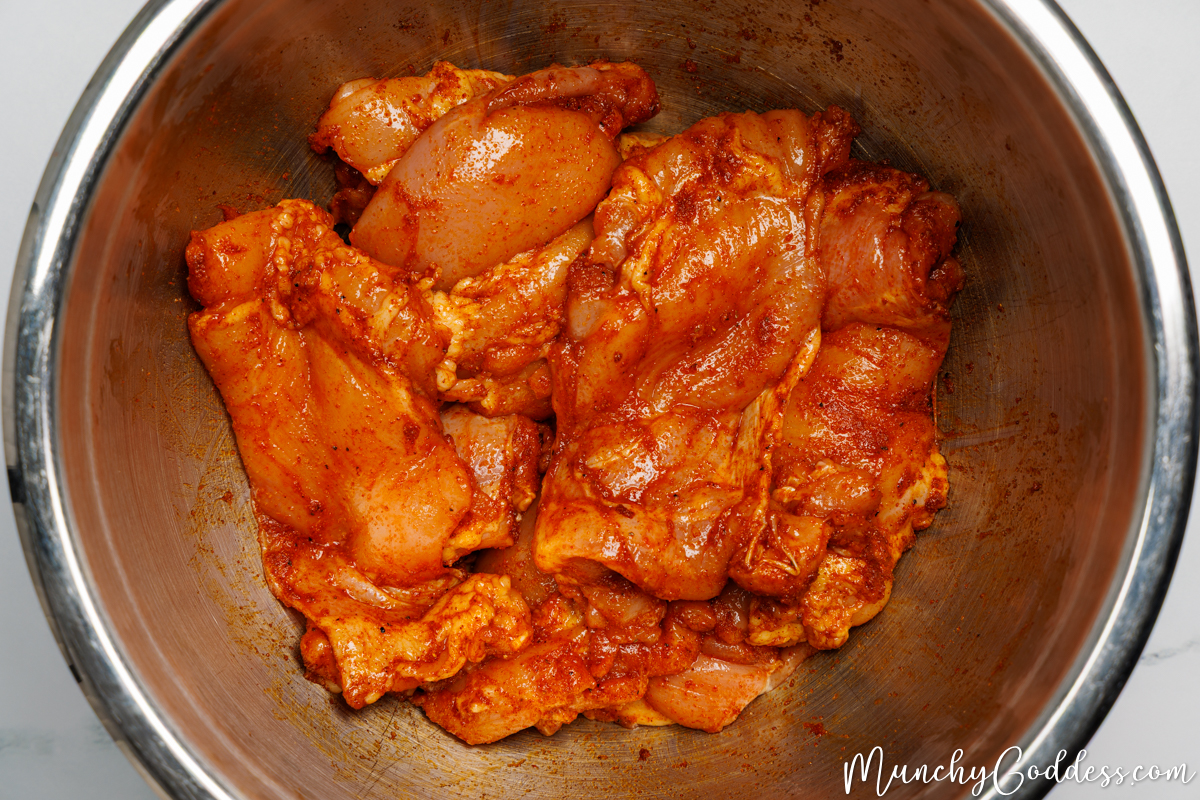 Boneless skinless chicken thighs tossed in an olive oil seasoning mixture until well coated in a silver mixing bowl.