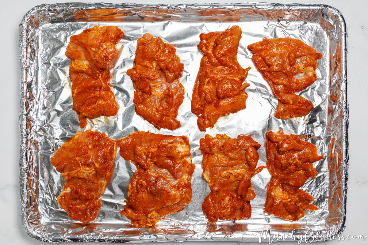 Boneless skinless chicken thighs coated in an olive oil seasoning mixture arranged in a single layer smooth side down on a sheet pan line with foil.