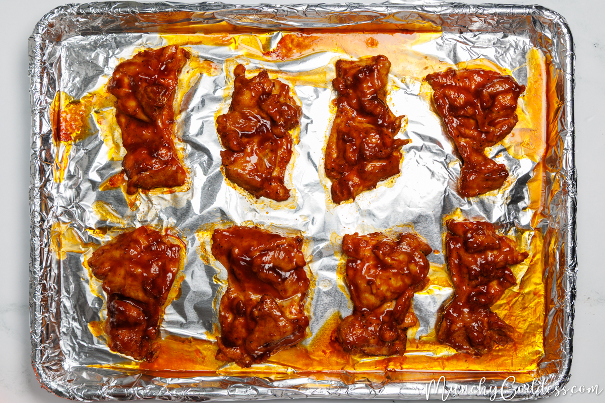Boneless skinless chicken thighs coated with BBQ sauce smooth side down on a sheet pan lined with foil.