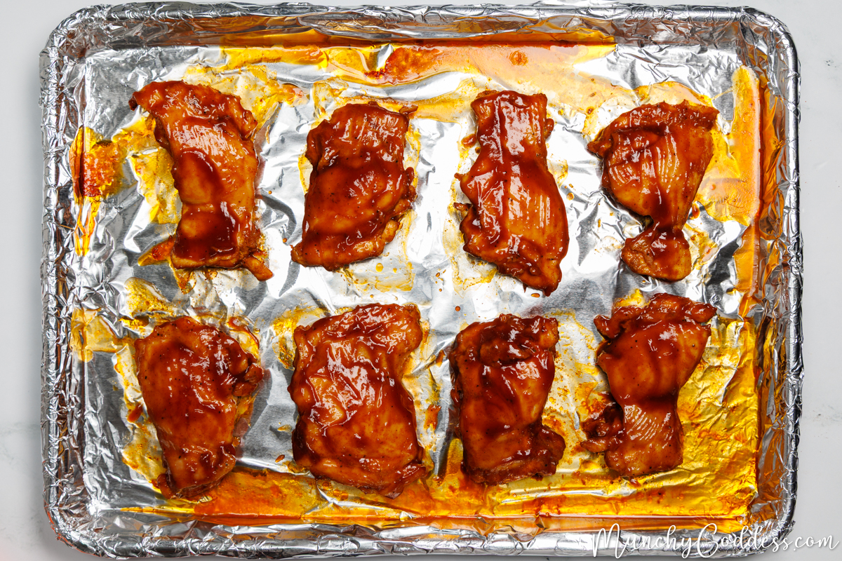 Boneless skinless chicken thighs coated with BBQ sauce smooth side up on a sheet pan lined with foil.