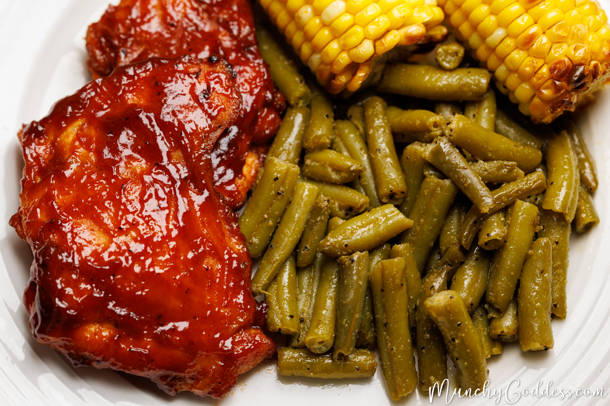 Baked BBQ chicken thighs with seasoned green beans and corn on the cob on an off-white plate.