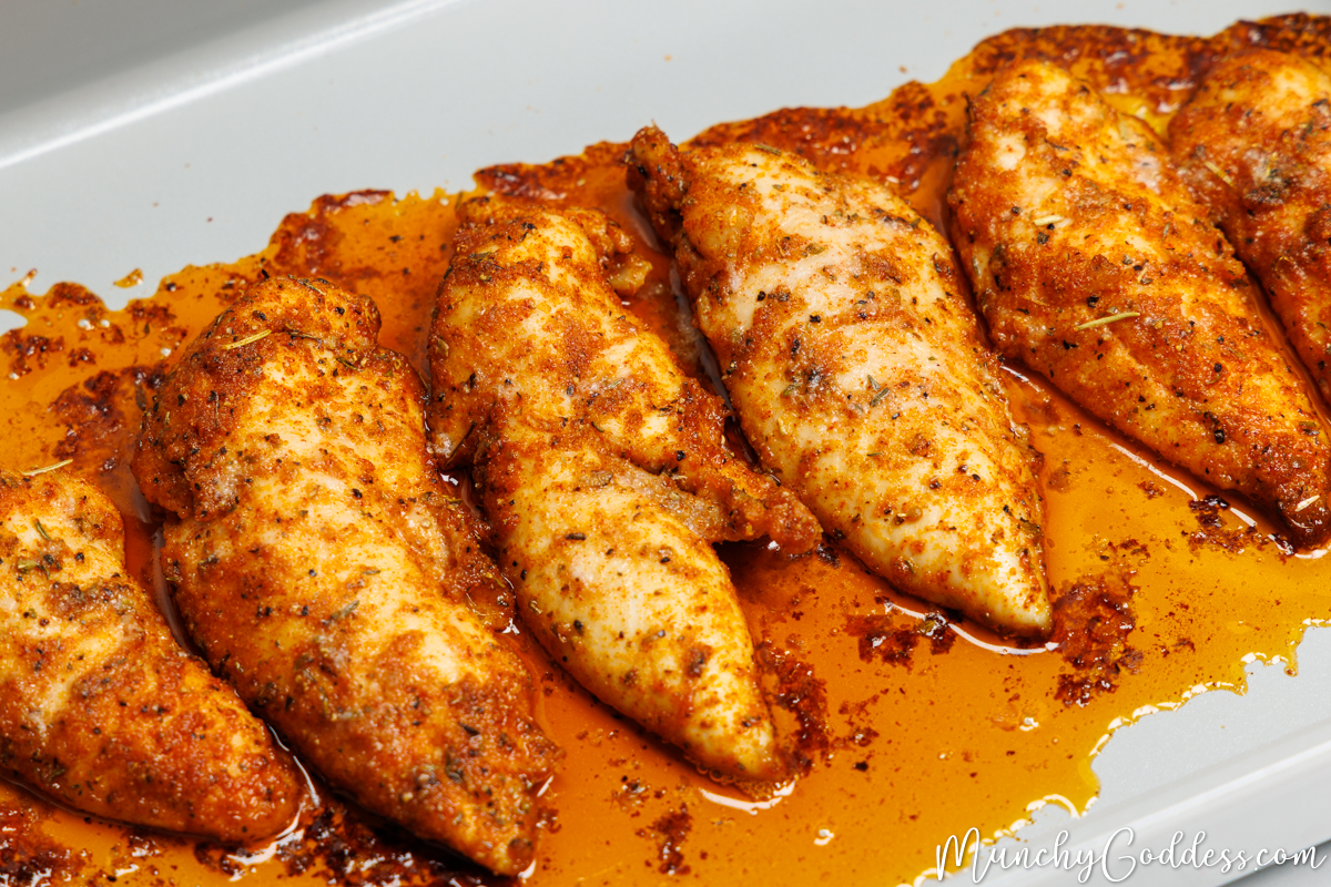 Baked chicken tenderloins in baked juices on light gray baking sheet.
