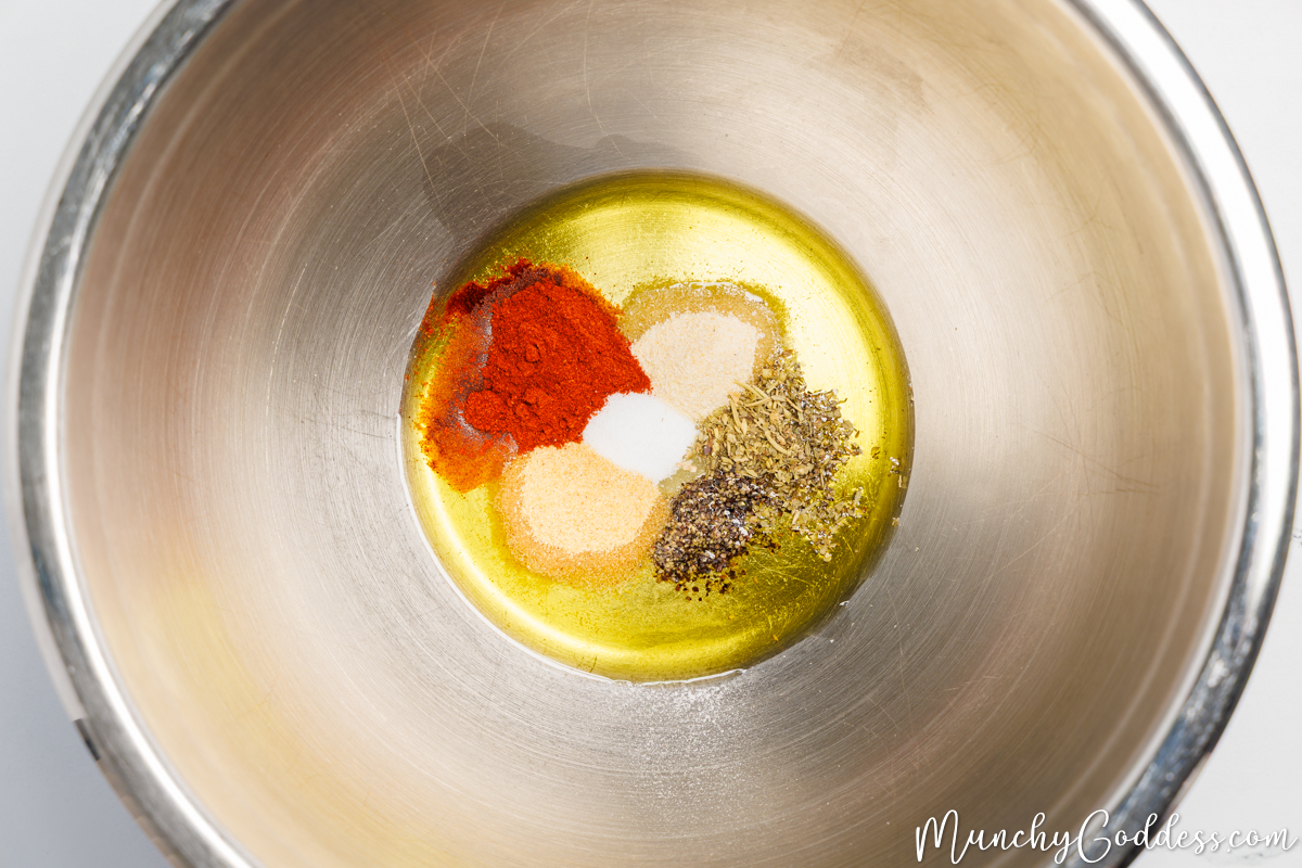 Olive oil, Italian seasoning, smoked paprika, salt, ground black pepper, garlic powder, and onion powder in a silver mixing bowl.