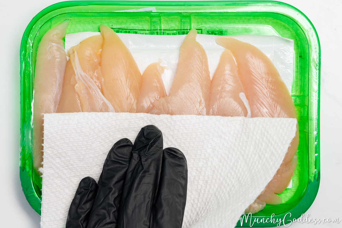 Chicken tenderloins in a green container being patted dry with a paper towel.