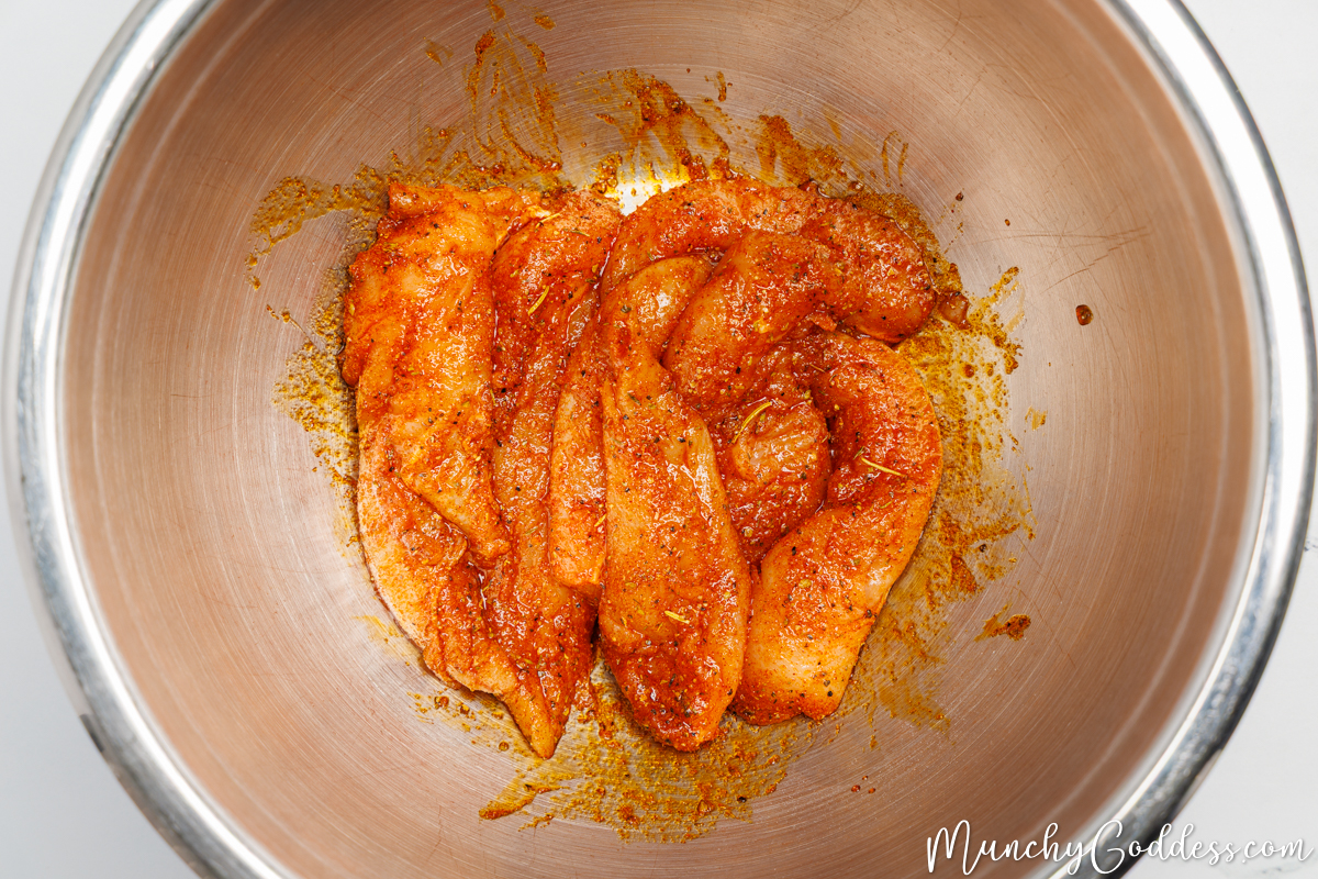 Chicken tenderloins well coated with an olive oil seasoning mixture in a silver mixing bowl.