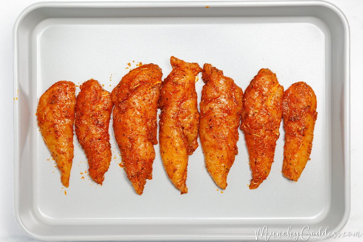 Chicken tenderloins coated in an olive oil seasoning mixture and arranged in a single layer in a light gray baking dish.