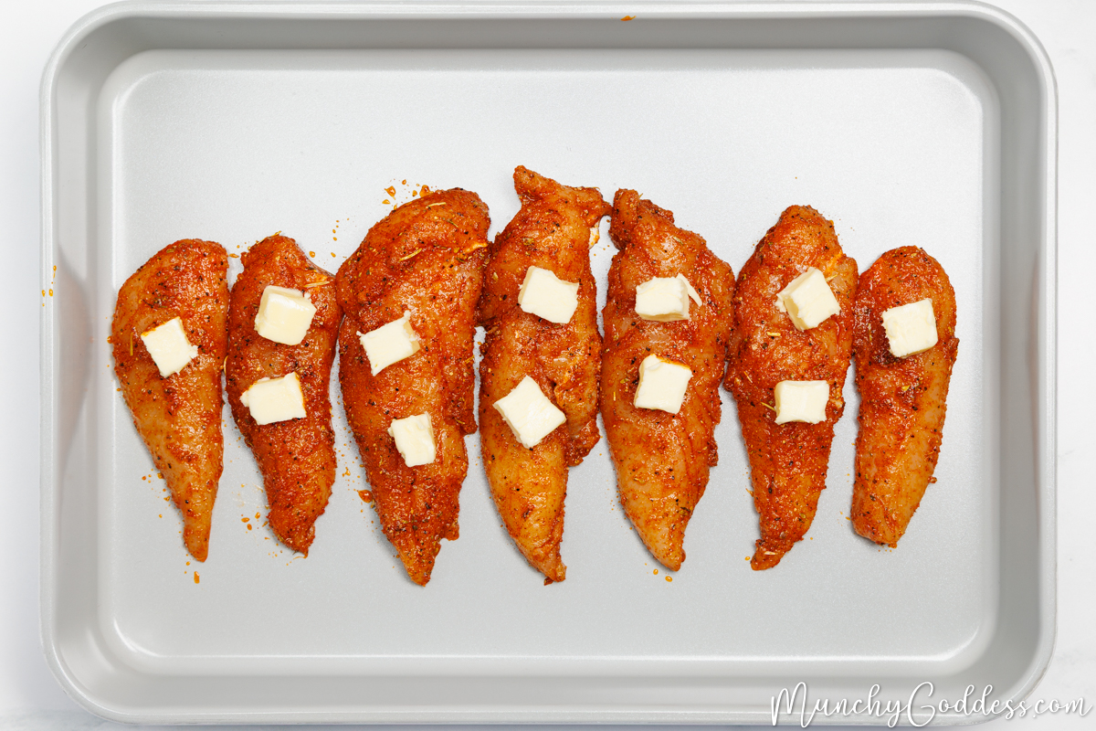 Chicken tenderloins coated in an olive oil seasoning mixture topped with pieces of butter and arranged in a single layer in a light gray baking dish.