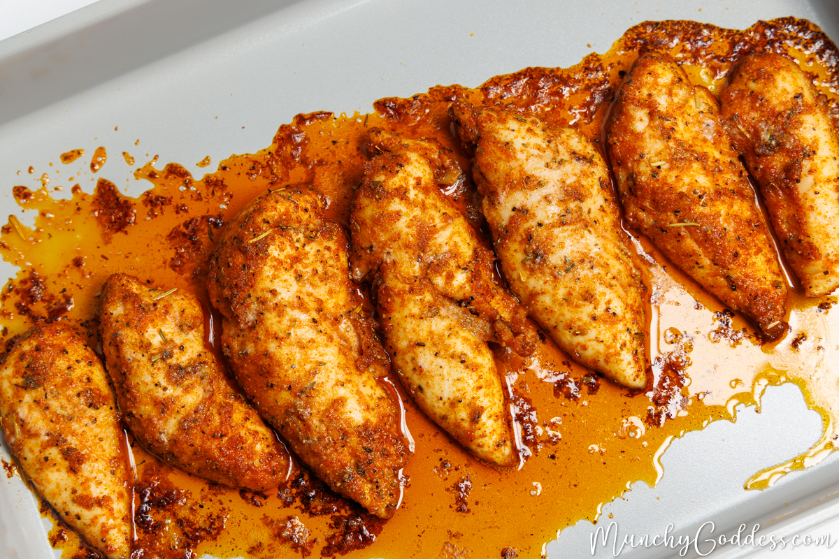 Seasoned and baked chicken tenderloins in glistening juices on light gray baking sheet.