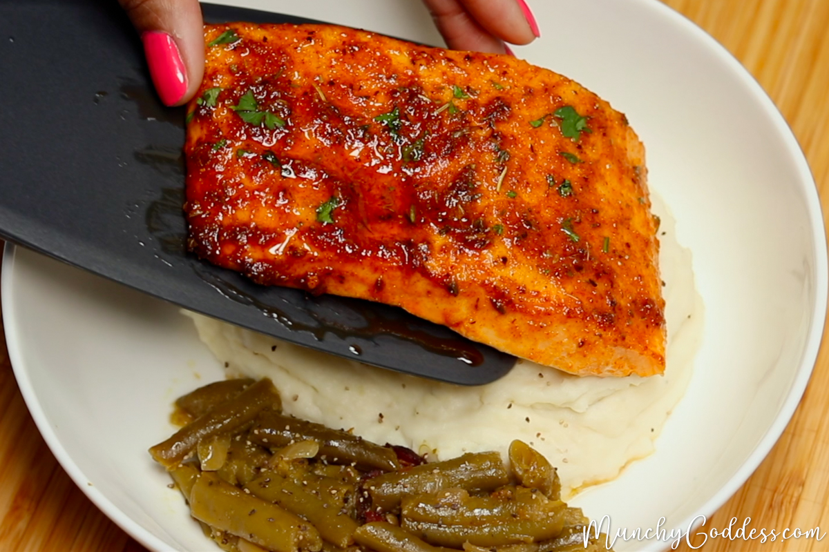 Salmon fillet sliding off a black silicone spatula onto a bed of mashed potatoes next to a side of green beans in an off-white bowl.