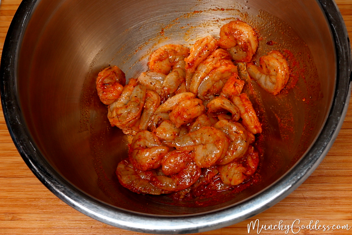 Raw jumbo shrimp well coated in a melted butter and olive oil seasoning mixture in a silver mixing bowl.