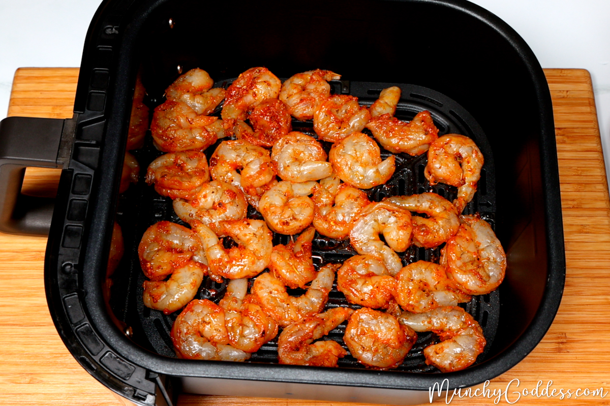 Seasoned raw jumbo shrimp added to a preheated black air fryer basket on a wood cutting board.